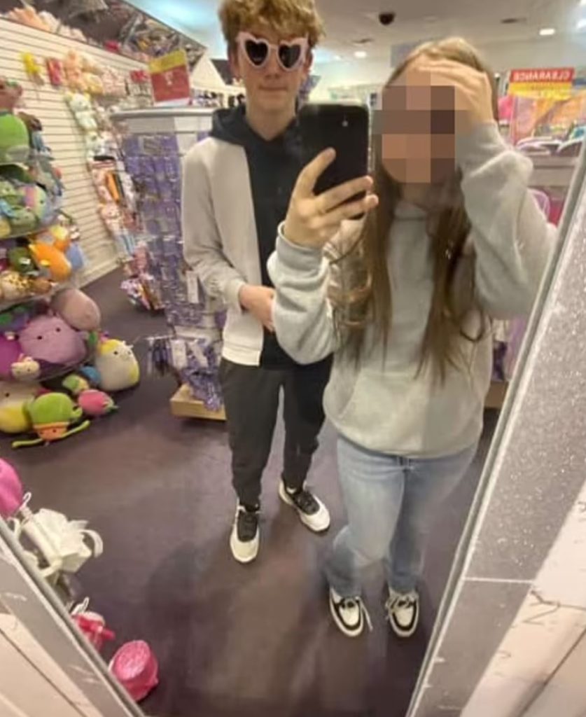 Young boy and girl pose in front of a mirror in a store full of plushies.