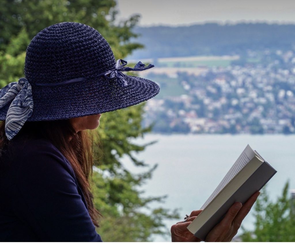Woman reading a book wearing a hat. Water and greenery in the background. 