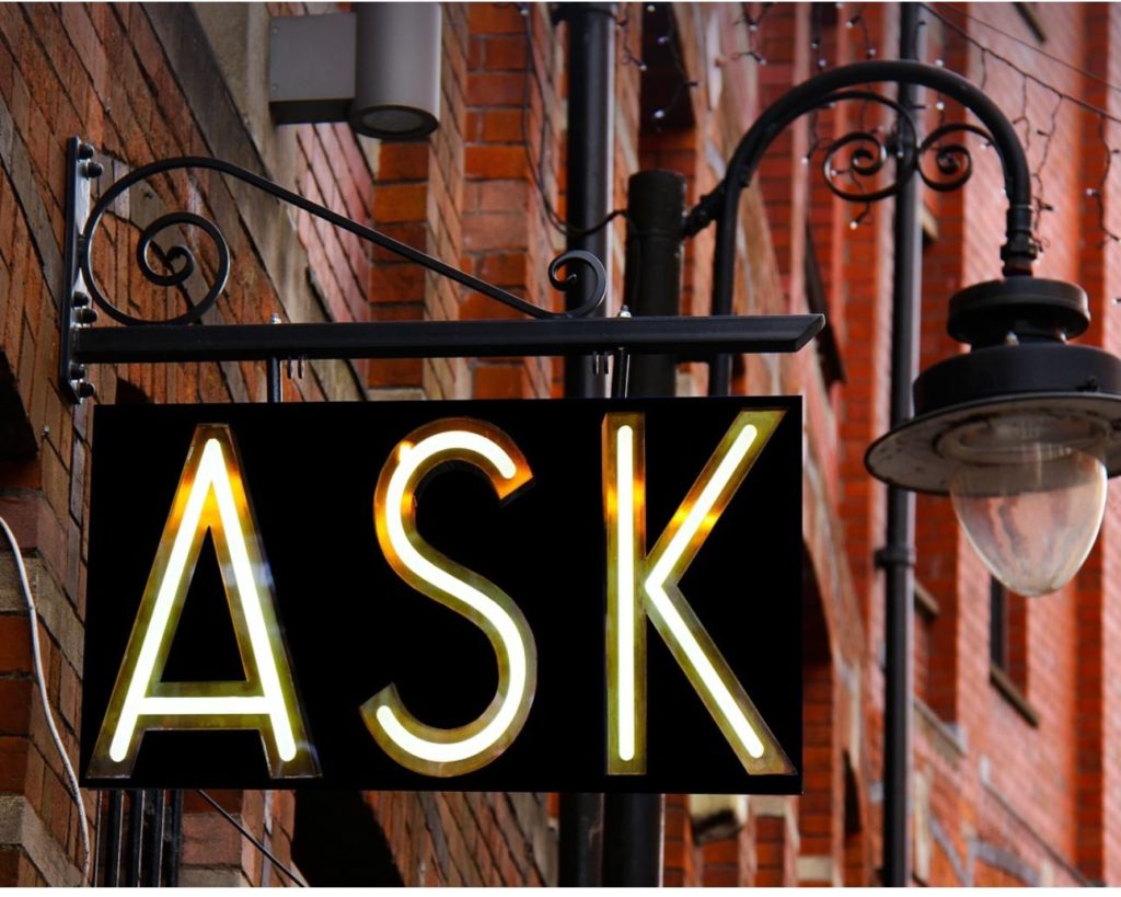 Sign that says ask with a streetlight and brick wall in the background.