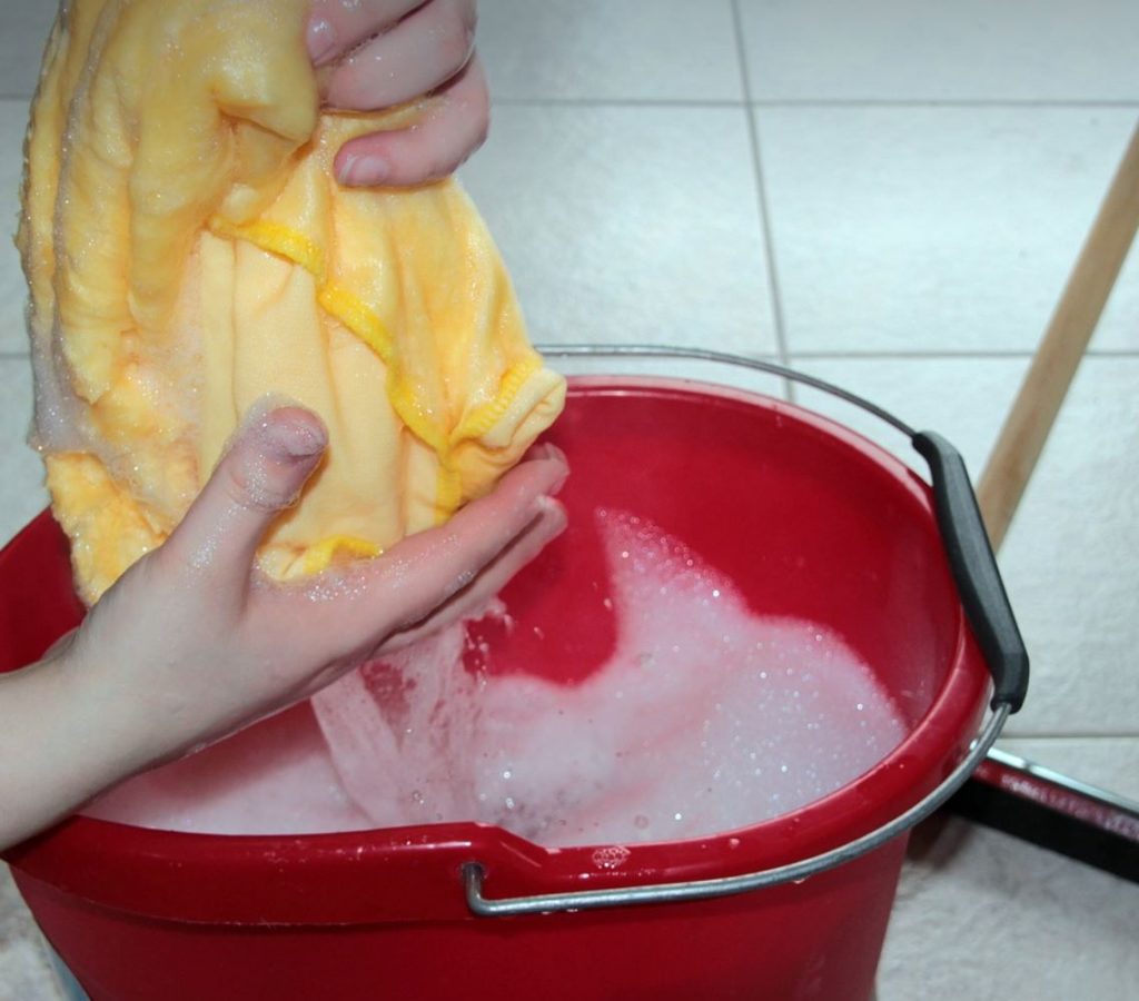 Red mop with soapy water and a person's hands squeezing out a yellow rag. 