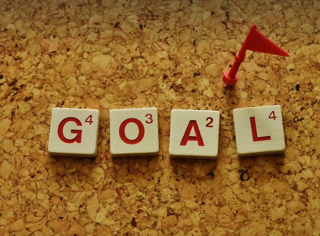 Scrabble letters spelling goal, on a wooden cork board with a red flag thumb tack. 
