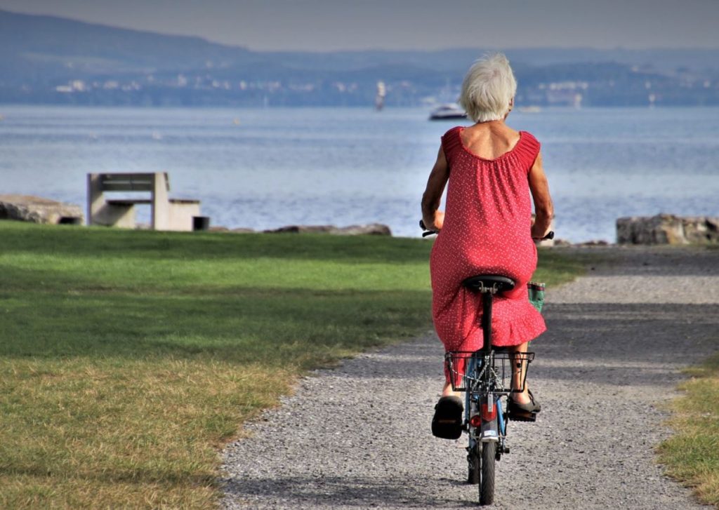 Older woman wearing red dress and riding a bike. Mountains, oceans, and beach towns in the background. 