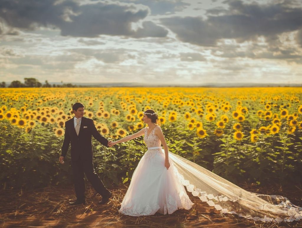 Bride and Groom holding hands in front of a field of sunflowers with a cloudy sky. 