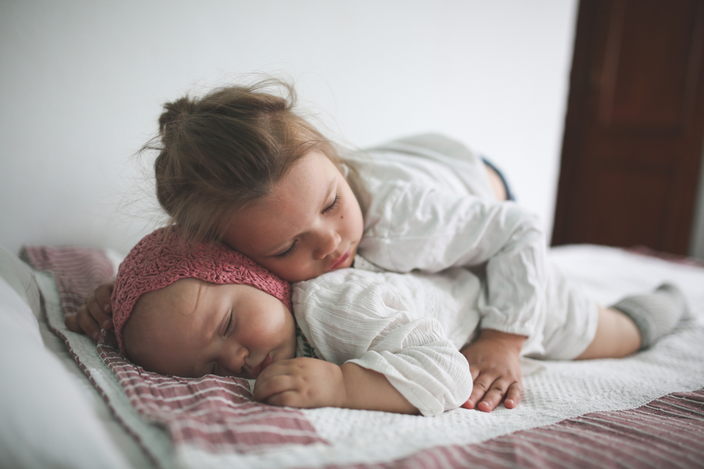 Toddler Girl Hugs a Sleeper Cute little baby in a crocheted cap on her stomach on a bed
