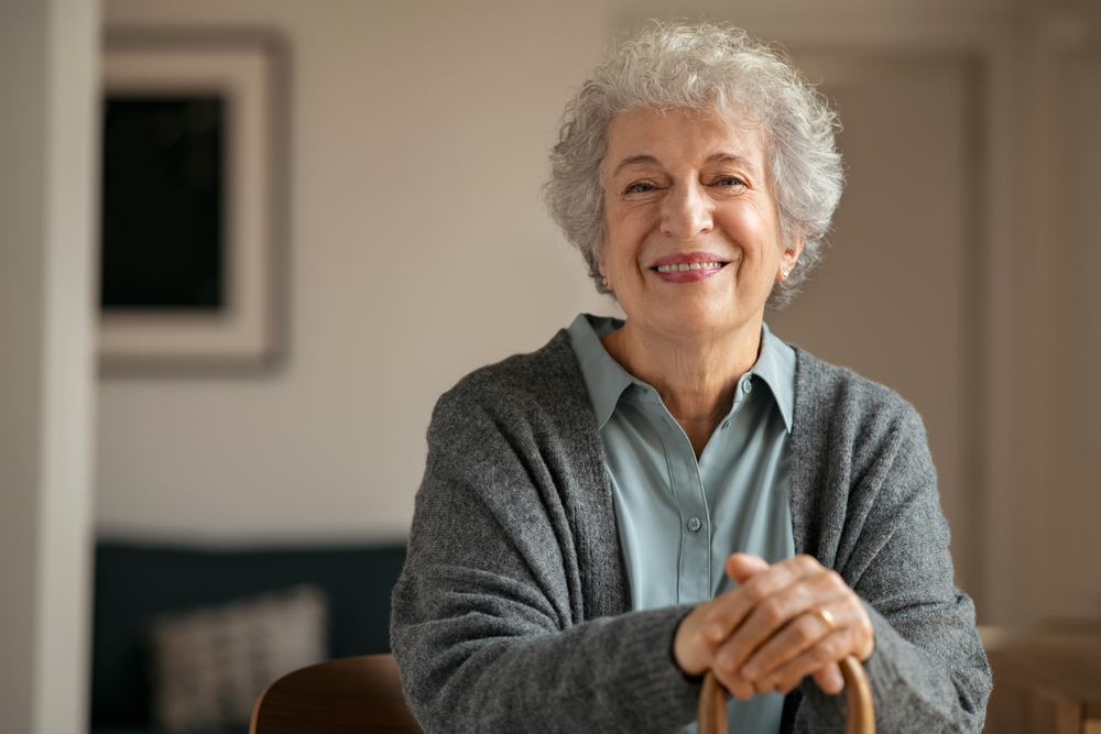 Portrait of smiling mature woman holding walking stick and sitting on chair at home. Portrait of happy senior woman under quarantine during covid-19 pandemic smiling while looking at camera.
