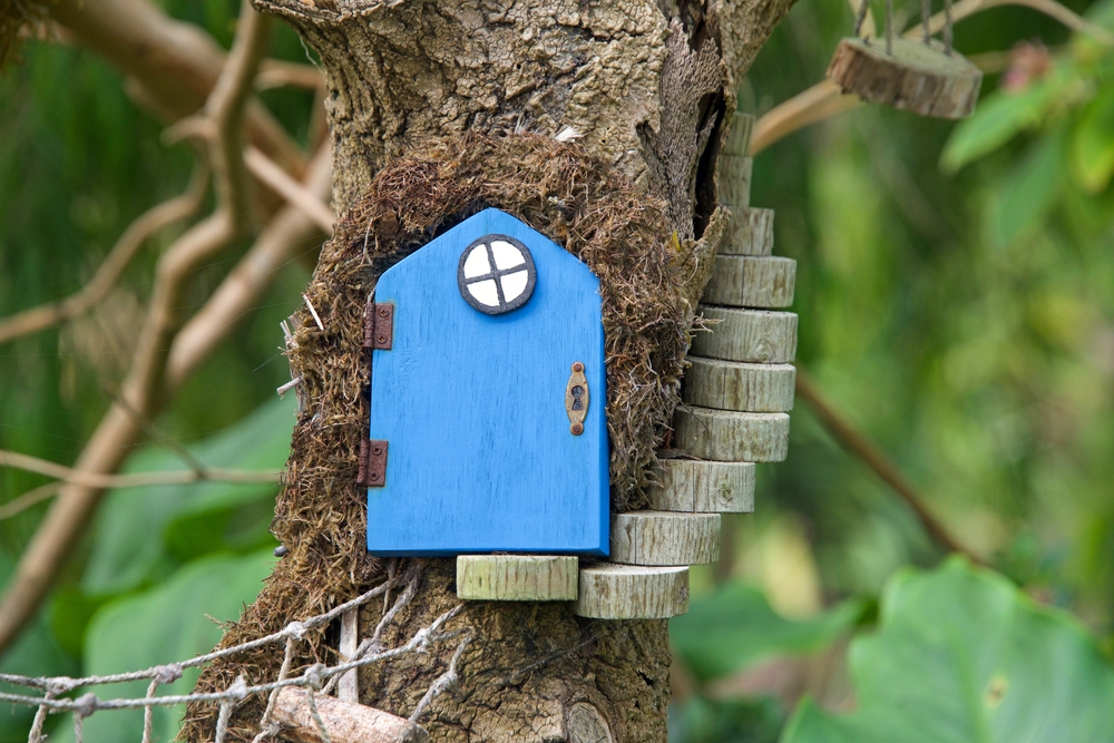 Close up of a fairy door with little steps to it, green leaves in background. An urban art movement of tiny fairy doors hitting the curbs, trees, and public spaces on the Island of Alameda, CA.