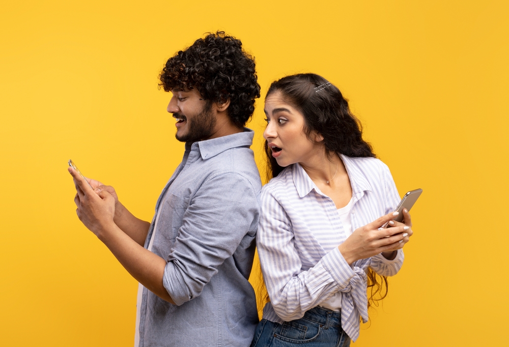 Shocked indian woman spying on her smiling boyfriend who using smartphone, chatting or scrolling social media news feed. Young couple standing back to back over yellow studio background