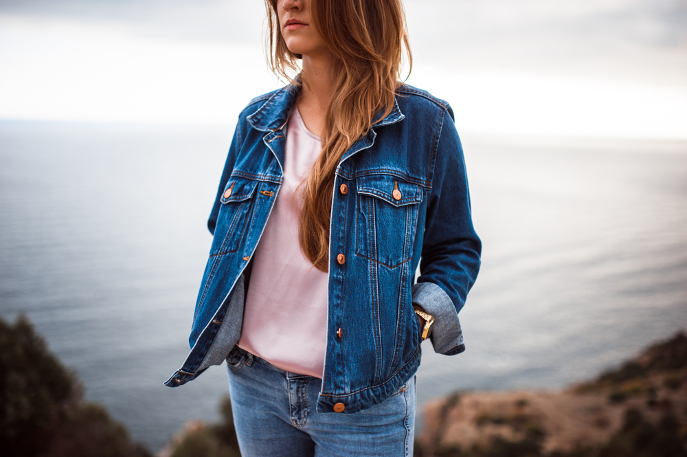 portrait of the attractive, slender, beautiful young Caucasian blonde girl in a jeans jacket. Smiling girl enjoys fine warm sunner weather highly in mountains against the sea