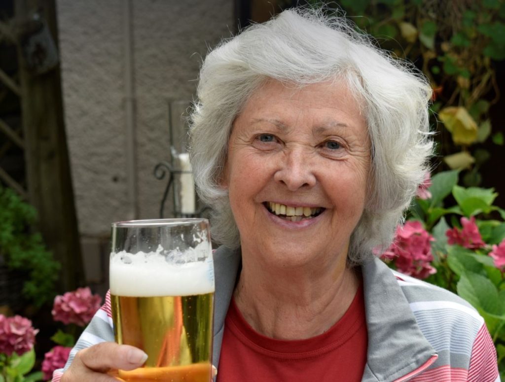 Older woman holding a beer. pink flowers and green leaves in the background. 