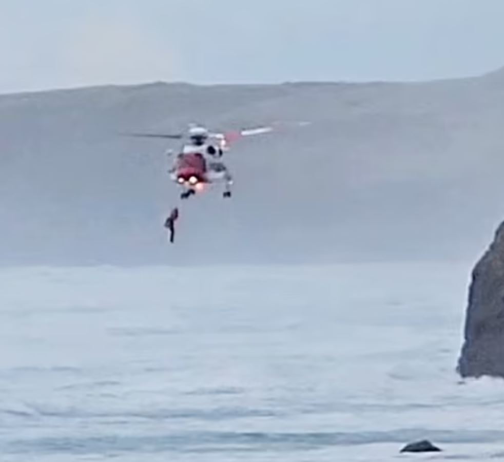 A man being rescued by a helicopter over the ocean. Fog and mountain in the background.