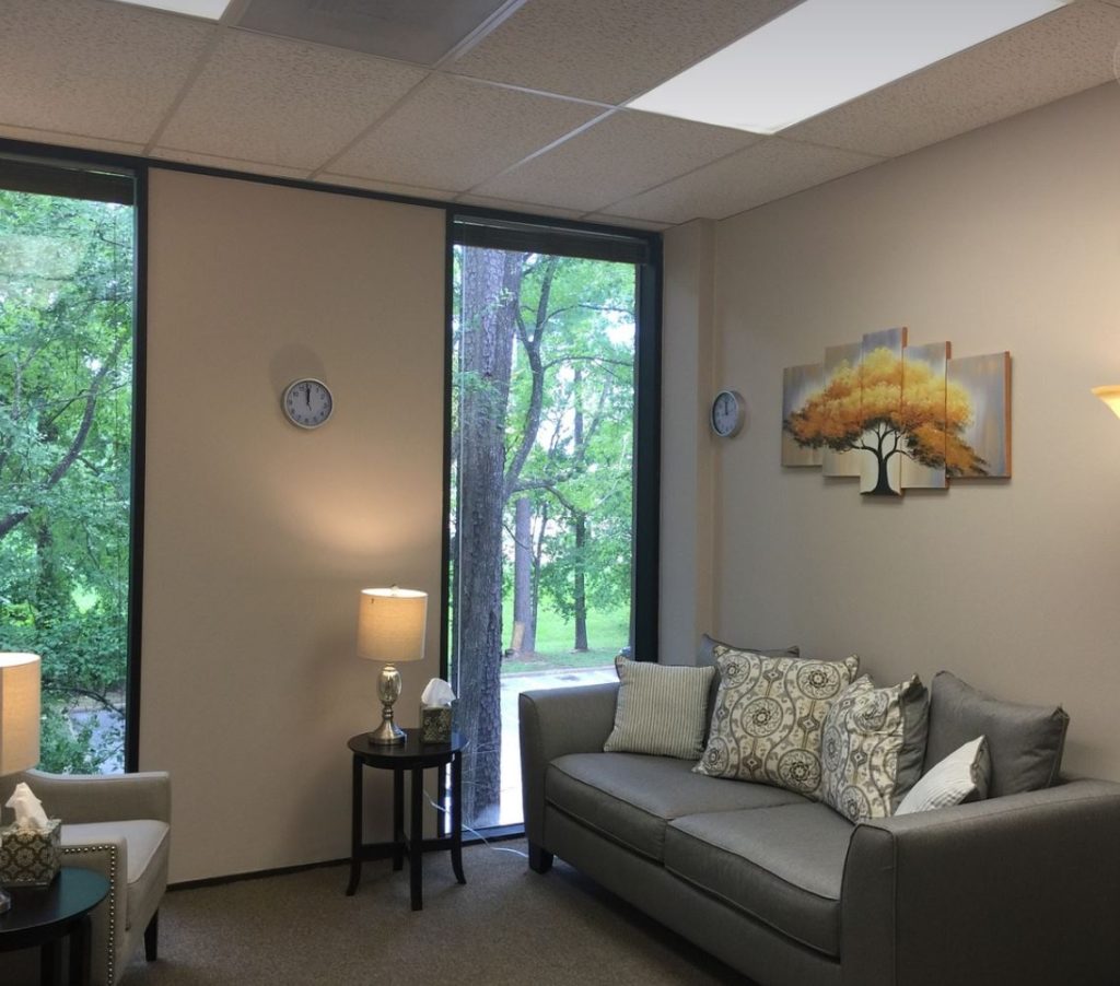 Therapist's office with couch and chair, pillows, windows, and side tables with lamps. Trees in the background.