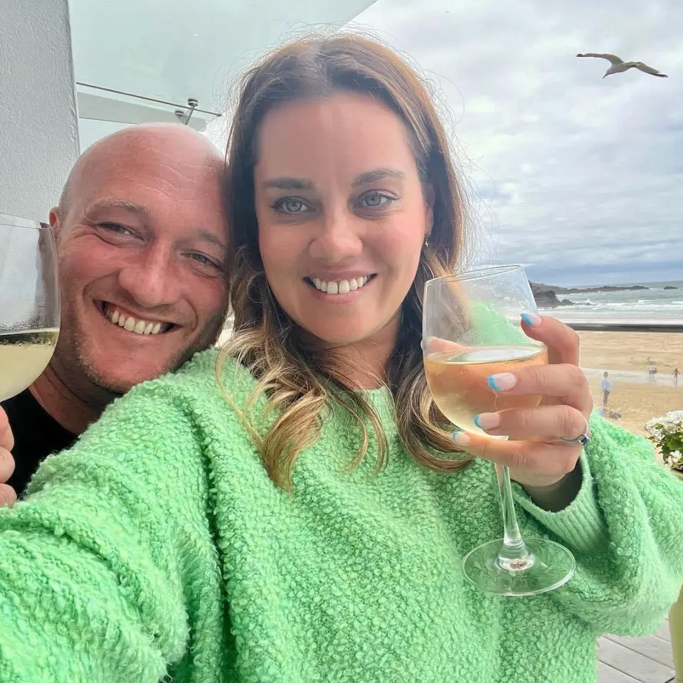 A man and woman smiling and holding a glass of wine. Beach in the background. 