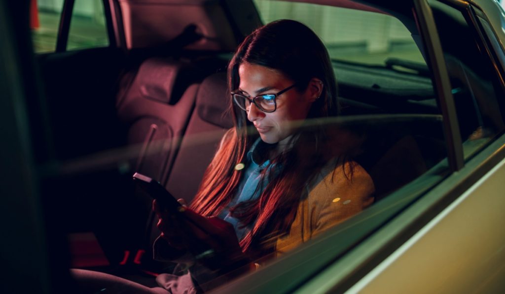 Businesswoman traveling with car on a business trip during night while sitting in a backseat and using a smartphone. Female using mobile phone to send email or messages. View trough car window.