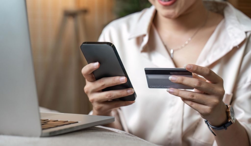 Mobile Shopping. Cheerful Asian Woman Using Smartphone Shopping Online Holding Credit Card Making Payment Sitting At Sofa At Home. Internet Banking Application And E-Commerce