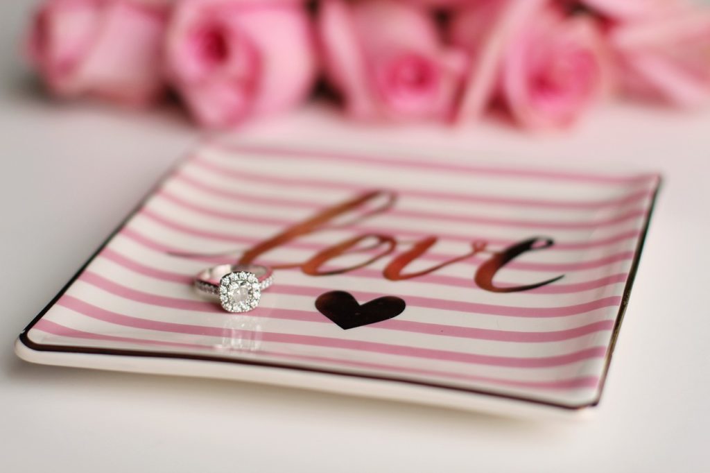 An engagement ring on a pink and white plate that says love in gold with a heart. Blurred pink roses and white background. 