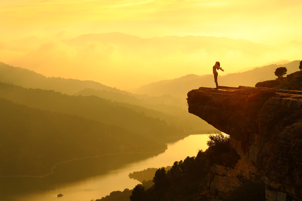 Woman silhouette breathing fresh air in the top of a cliff in the mountain at sunset with a beautiful landscape in the background