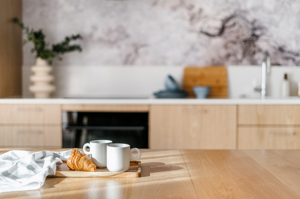 dish towel, fresh croissant and ceramic cups of tea on bamboo tray on wooden tabletop with sun light on kitchen background interior, breakfast concept