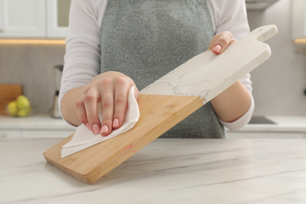 Woman wiping cutting board with paper napkin at white table, closeup