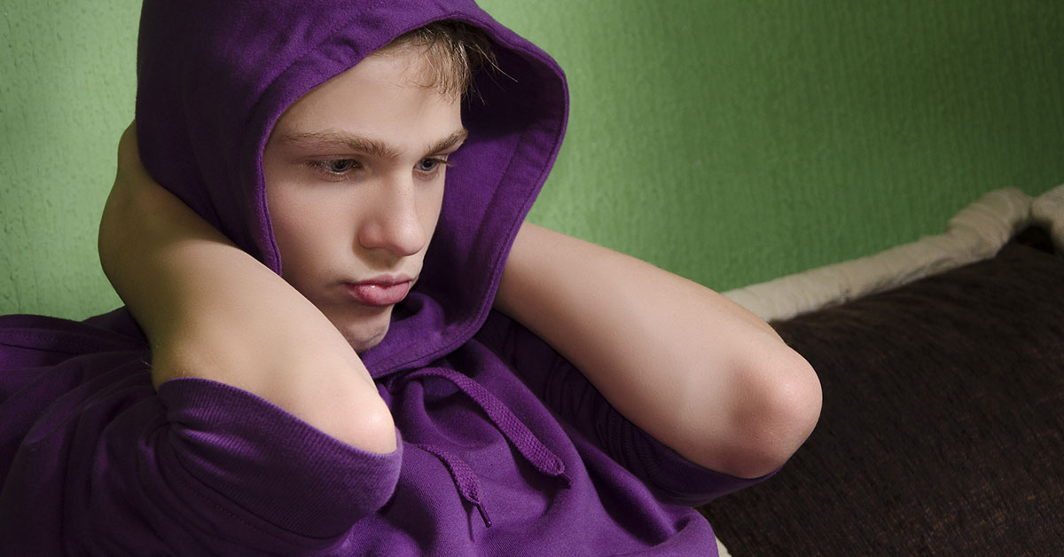 Depressed teenager with arms around his neck and hood on head, looking down.