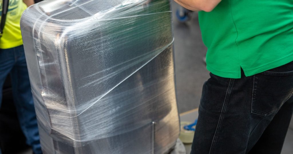 Worker wrapping suitcase with transparent protection film atairport deparure terminal before flight. Luggage protection and safety service