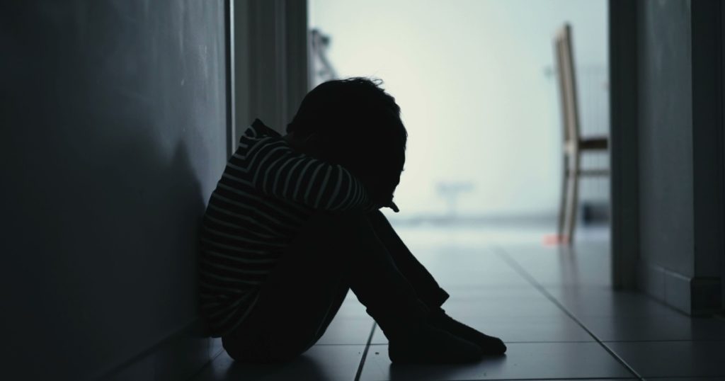 Sad child suffering from depression sitting alone in corridor feeling loneliness. Scared fearful small boy covering face in silhouette at home