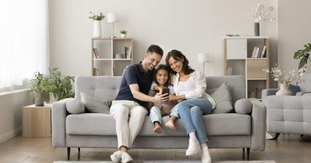 Positive young mom, dad and sweet girl kid taking family selfie on cozy comfortable couch, having fun with digital gadget in cozy stylish home interior, using smartphone for selfie together