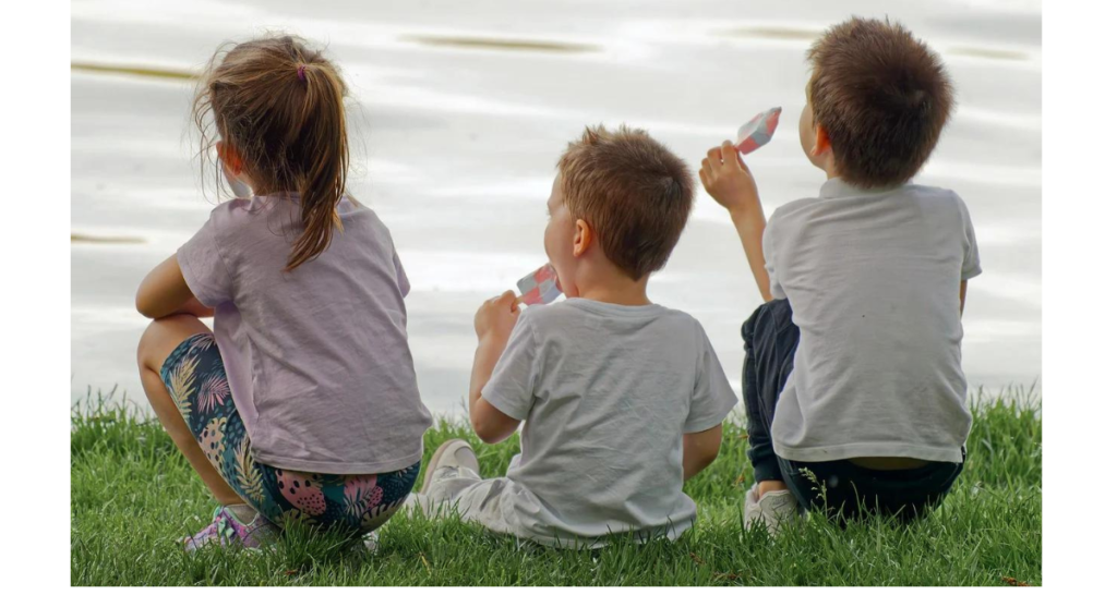 Three children eating ice cream and sitting in the grass with their backs to the camera. Water in the background. 