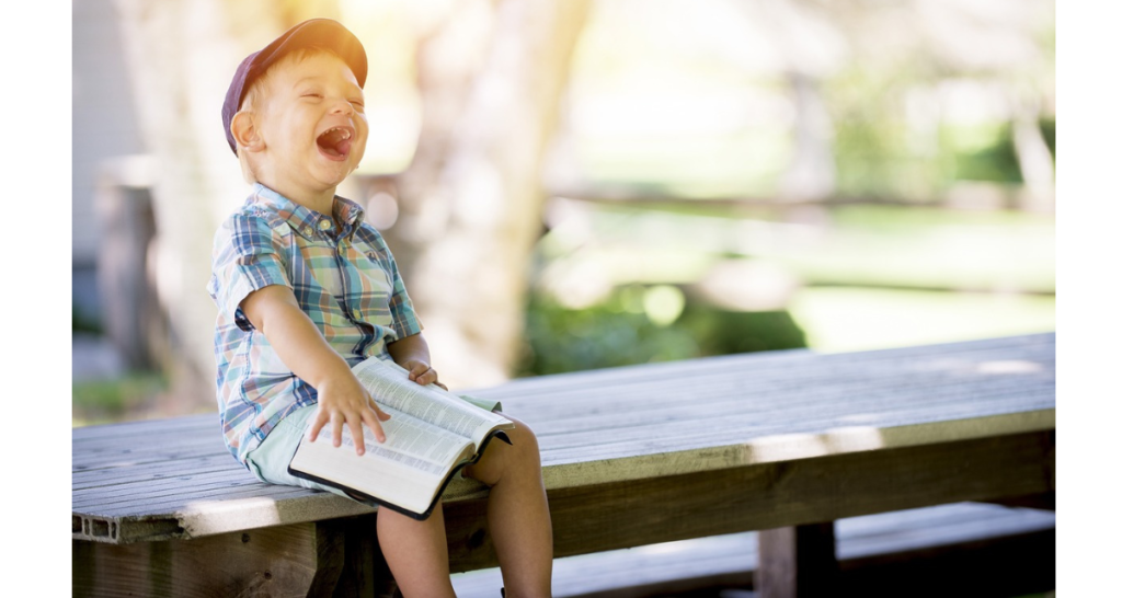 Boy reading a book and laughing at a park. Blurred greenery and trees in the background. 