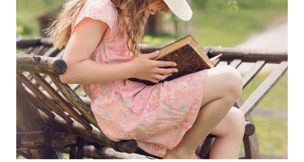Girl in a pink floral dress sitting on wooden structure reading a book. Grass and a path in the background. 
