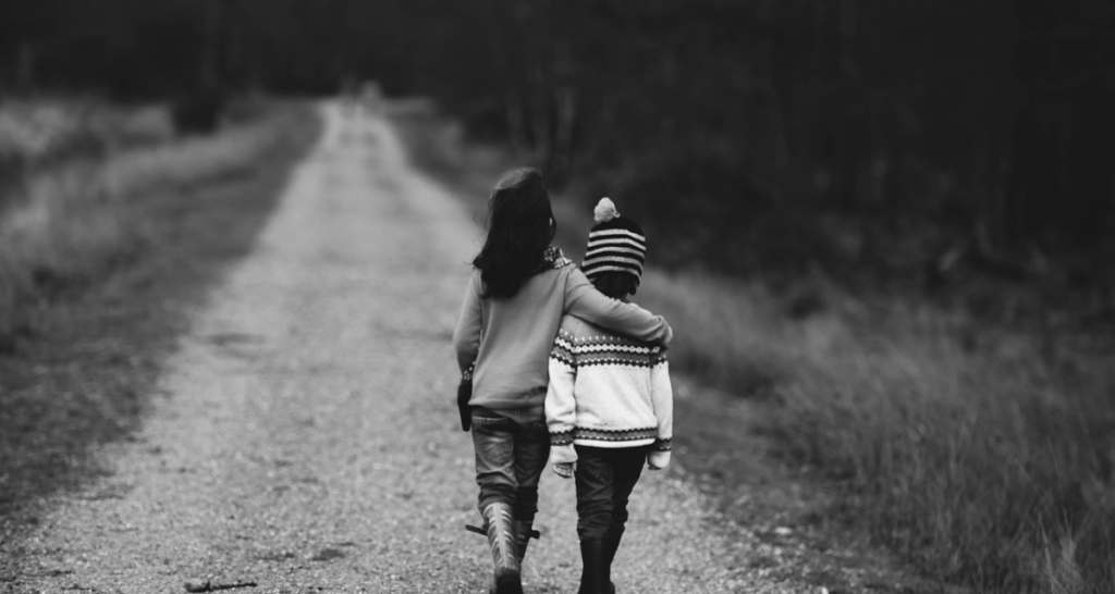 A black and white photo of two children walking together in an embrace down a road. Plants and trees in the background. 