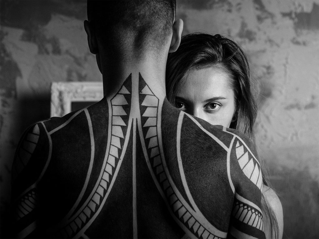Balck and white image of a woman looking over a man's shoulders. His tattooed back is toward the camera.