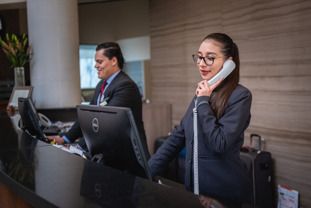 Receptionists answering phones at the front desk. Luggage and a wall in the background.