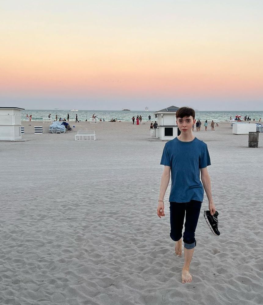 Young man standing bare foot on the beach. Sand, ocean, sunset, and people in the background.