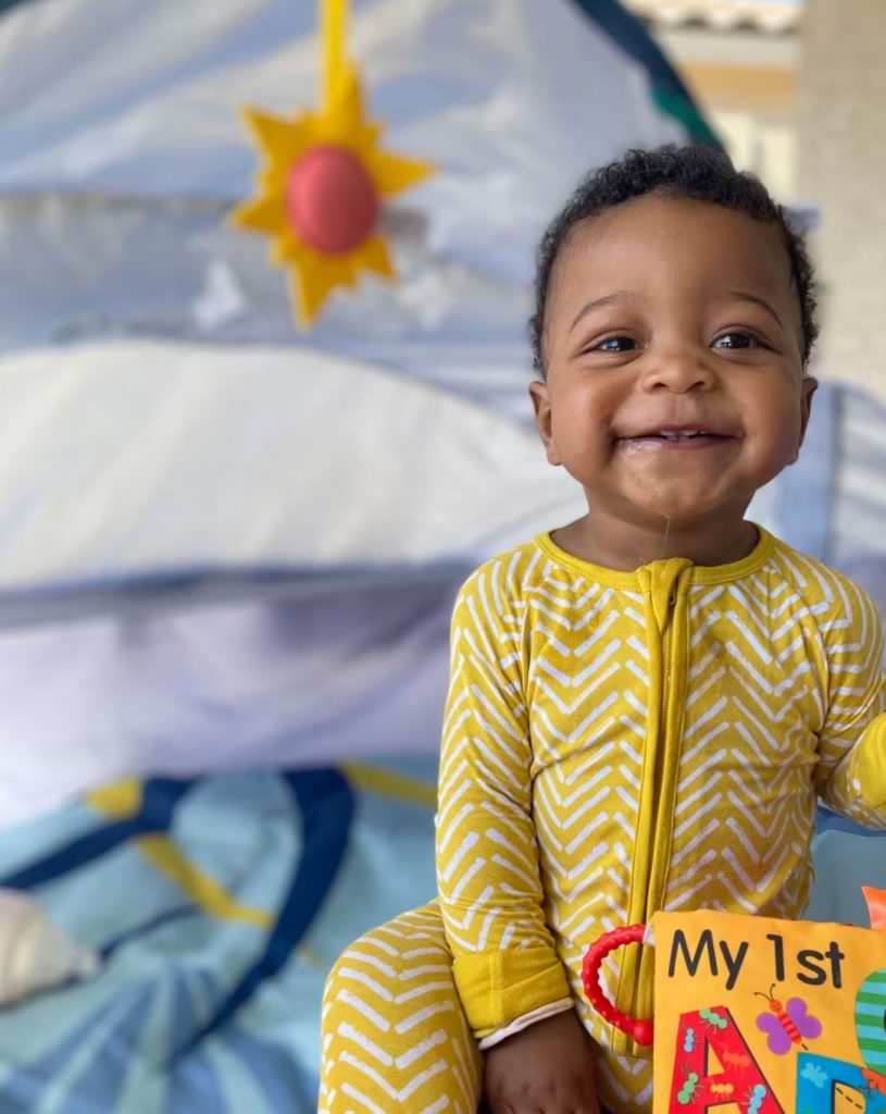 The 2024 Gerber Baby smiling in a yellow and white zigzagged striped onesie. Blurred baby play area in the background.