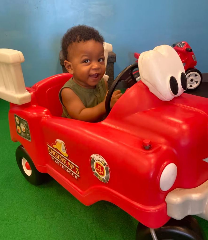 A little boy riding a red plastic car. Green and blue in the background.