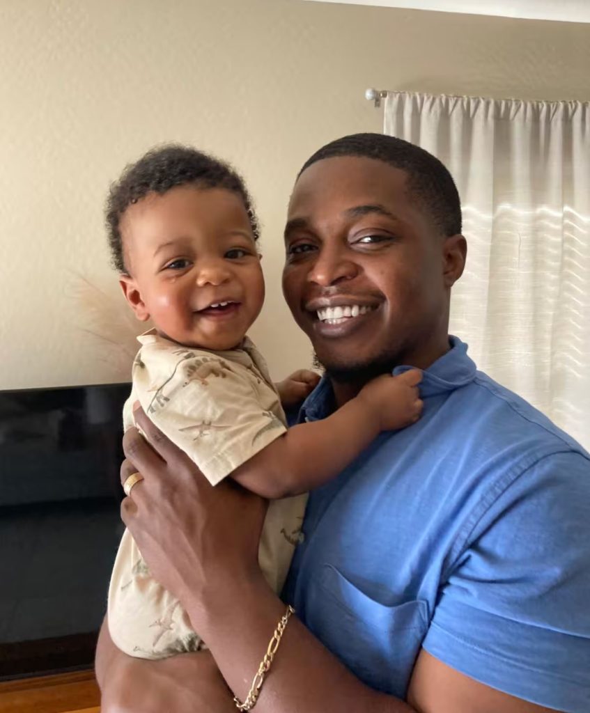 The Gerber Baby smiles with his dad. Curtains, a tv, and wall in the background.