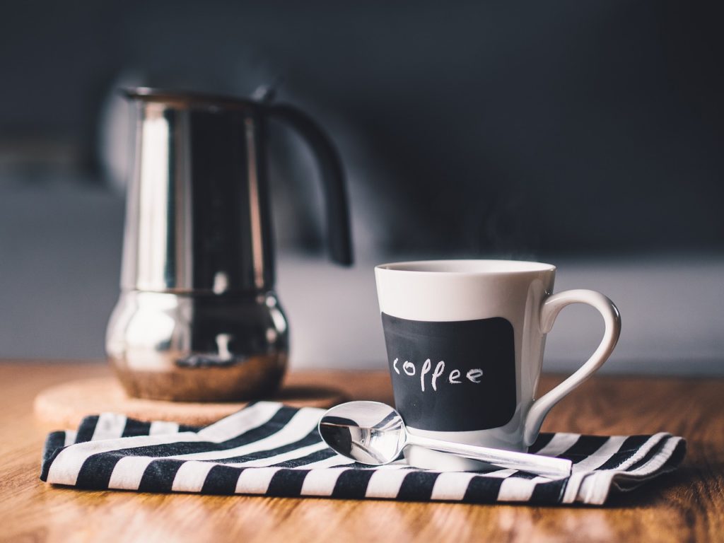 A coffee mug that says coffee with a spoon and coffee pot, Black and white striped towel and wooden table in the background.