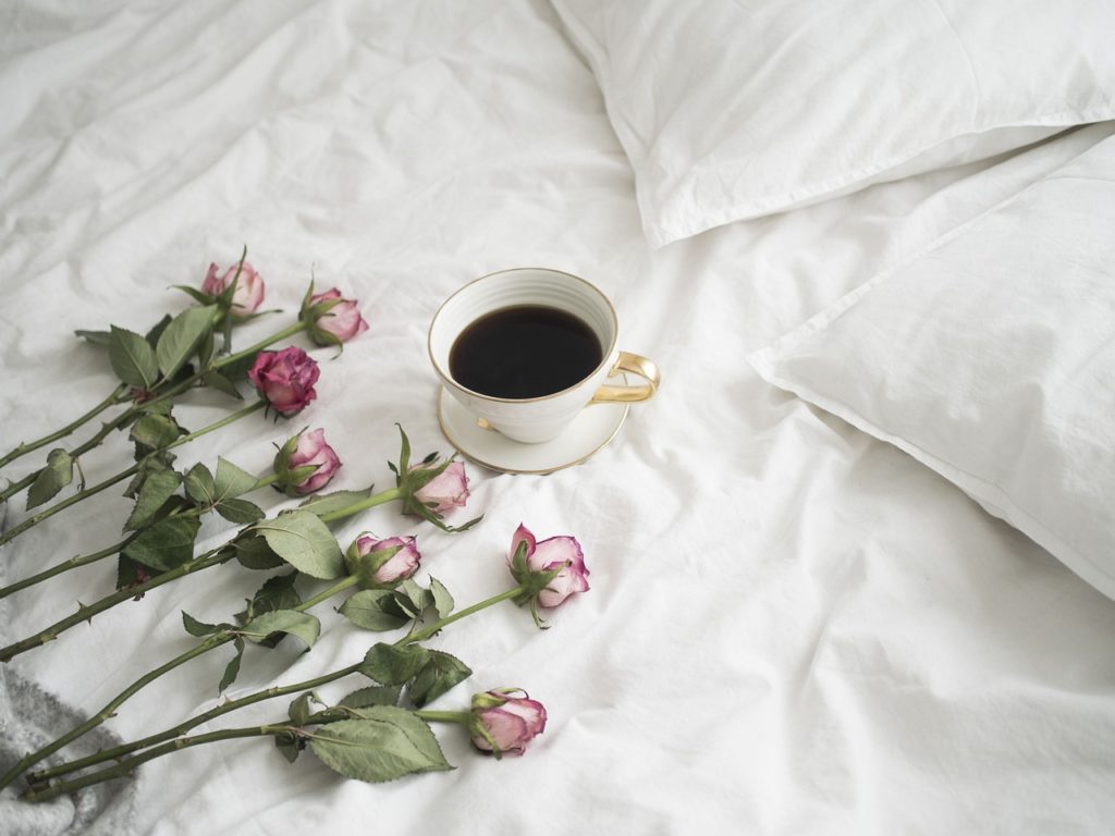 A hotel bed and pillows with a cup of coffee and pink roses.