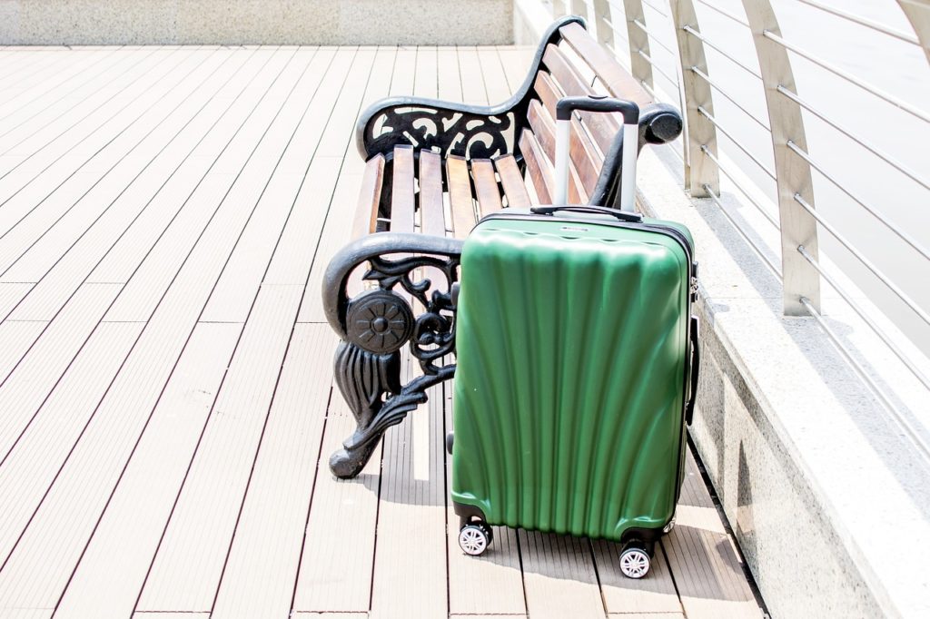 A green suitcase with wheels and a raised handle next to a bench. Wooden flooring and a guardrail in the background.