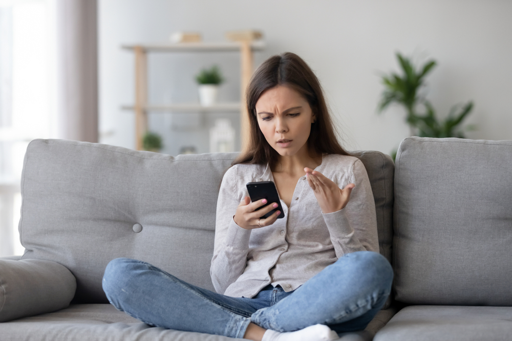 Indignant young woman sitting on couch in living room holding mobile phone having problems with digital device, feels angry and annoyed. Discharged broken not working and out of order gadget concept
