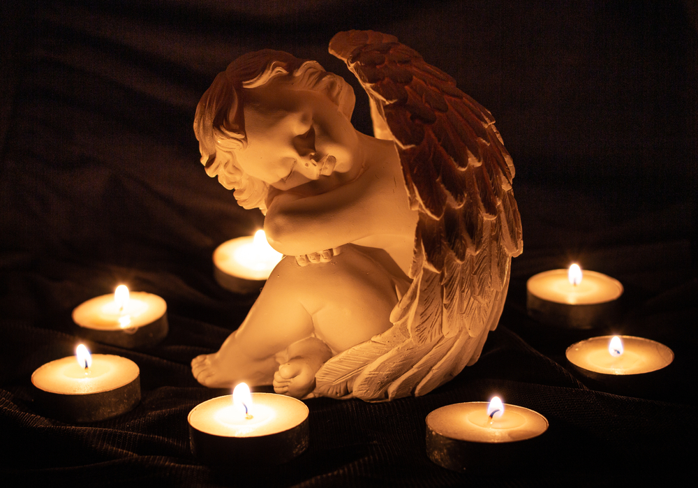 angel sculpture surrounded by burning candles; grief and loss memory of the deaths concept