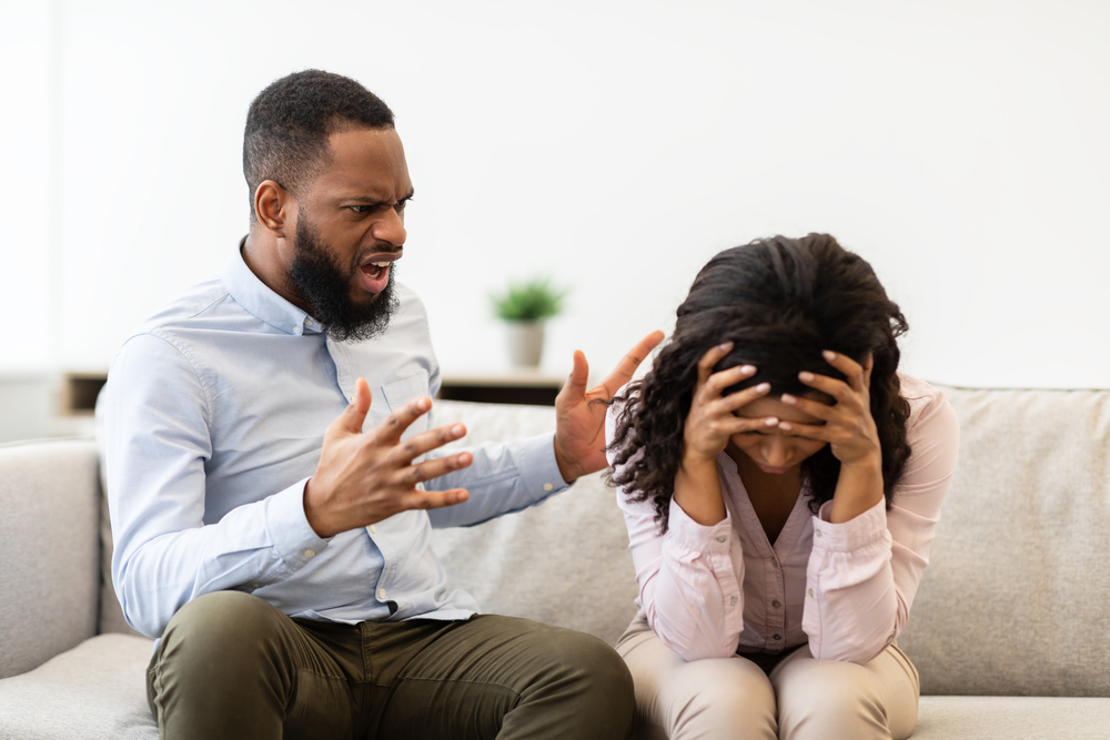 Breaking Up And Divorce Concept. Young married black couple having fight, guy yelling at crying lady, gesturing at home. Furious man shouting at his wife or girlfriend, sitting on sofa at living room