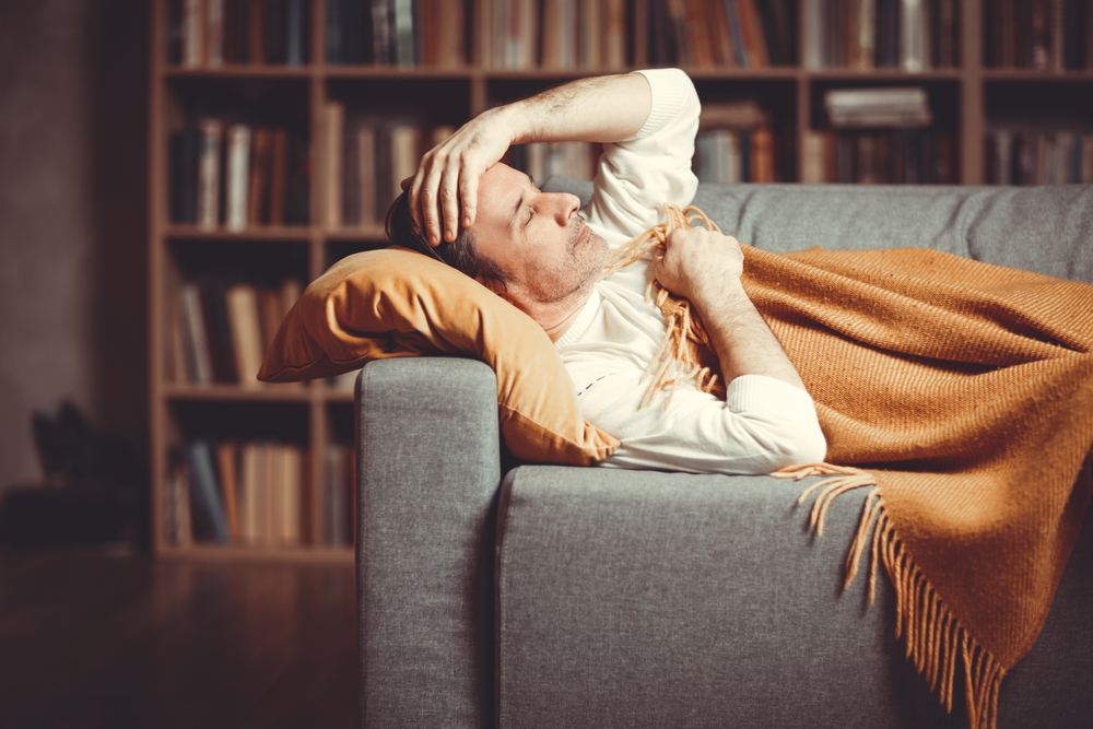 An adult man lies on the couch at home against the background of a bookcase with symptoms of the disease.