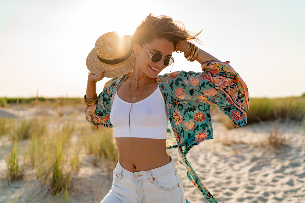 pretty attractive slim smiling woman on sunny beach in summer style fashion trend outfit happy, freedom, wearing white top, jeans and colorful printed tunic boho style chic and straw hat