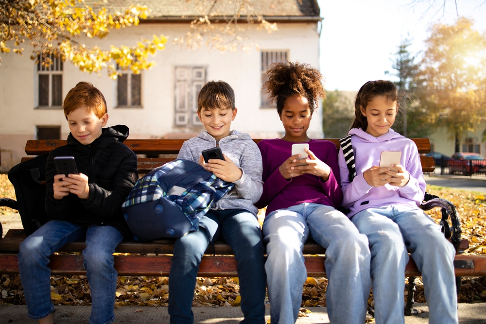 Group of multicultural children sitting on bench and using their smart phones surfing internet - technology concept.