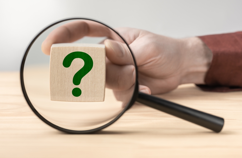 Hand holding question mark in front of magnifying glass. Search for an answer to question. Magnifying glass with question mark in focus on wooden cube. Research, searching or investigating something