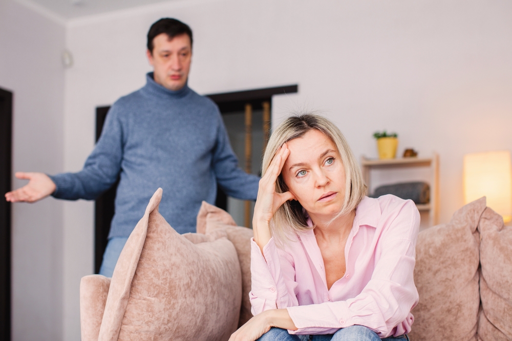 Sad middle-aged woman sits upset on the couch at home while his angry husband swears at her. Conflict in the family