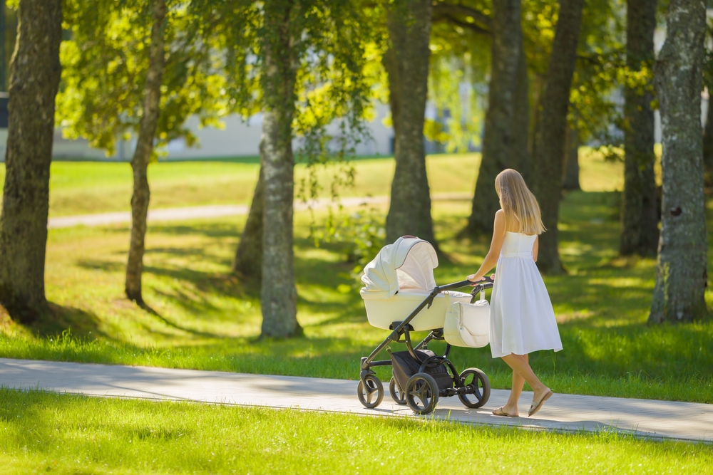 Young adult mother in dress pushing white baby stroller and walking on sidewalk at town park in warm sunny summer day. Spending time with infant and breathing fresh air. Enjoying stroll.