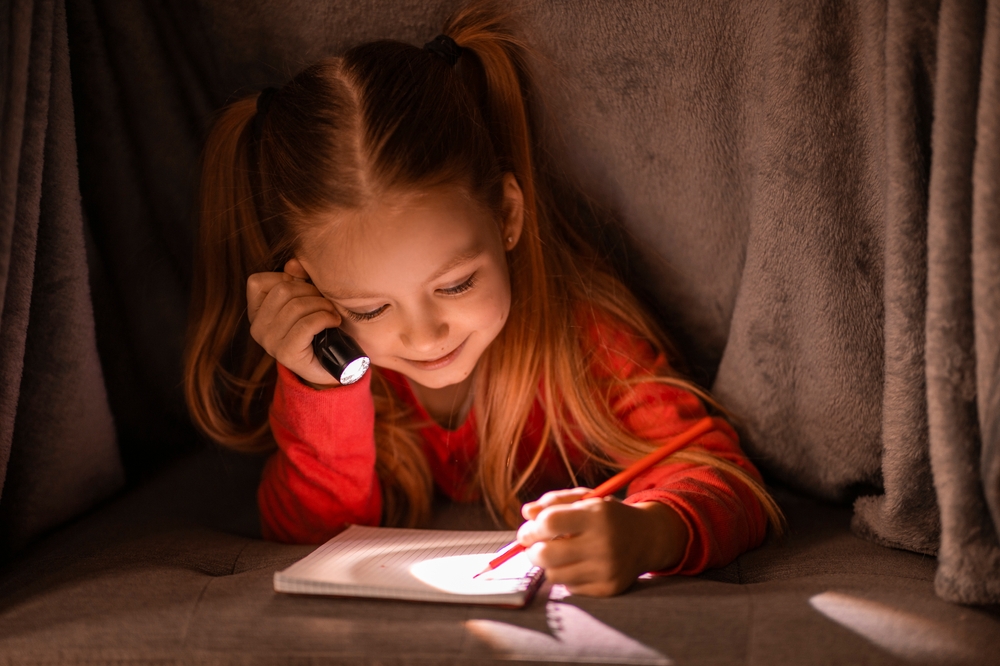 Cute little girl with flashlight hiding under blanket and writing in notebook, smiling preteen female child relaxing under covers and drawing in notepad, filling diary, using torch in the dark