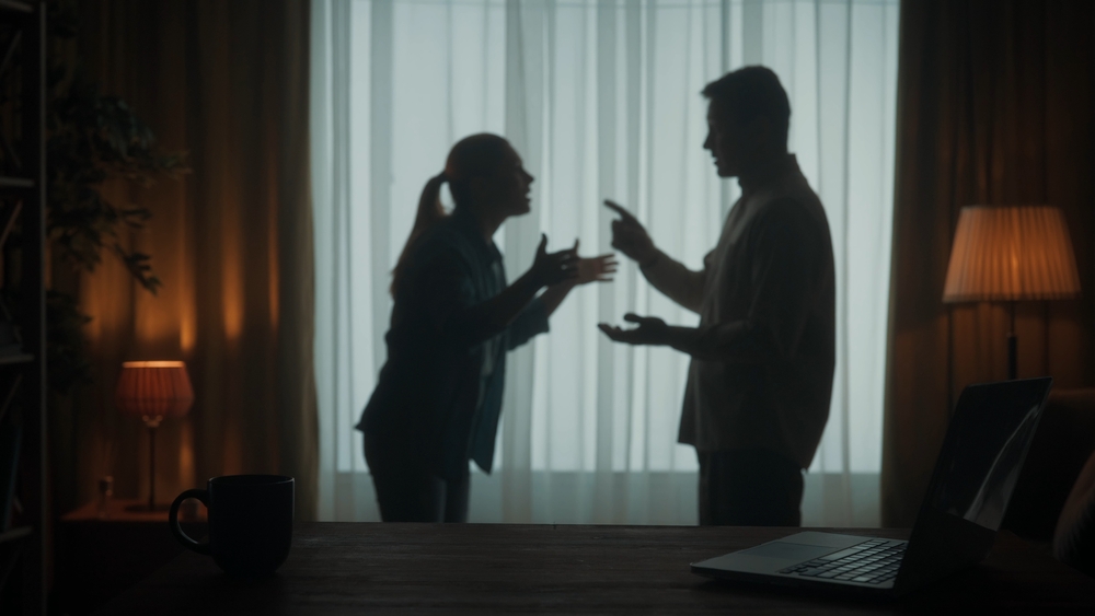 A man and a woman are arguing in the living room near the window. A silhouette of a couple arguing over the fact that the man spends too much time on the laptop.